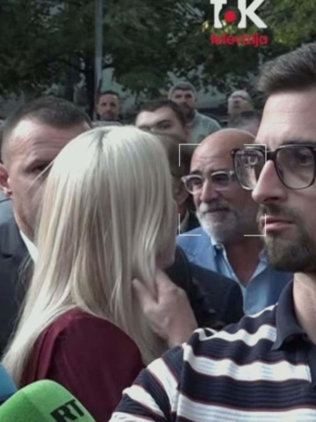 Video frame of a crowded outdoor scene with a reporter’s microphone in the foreground and a TV channel logo at the top