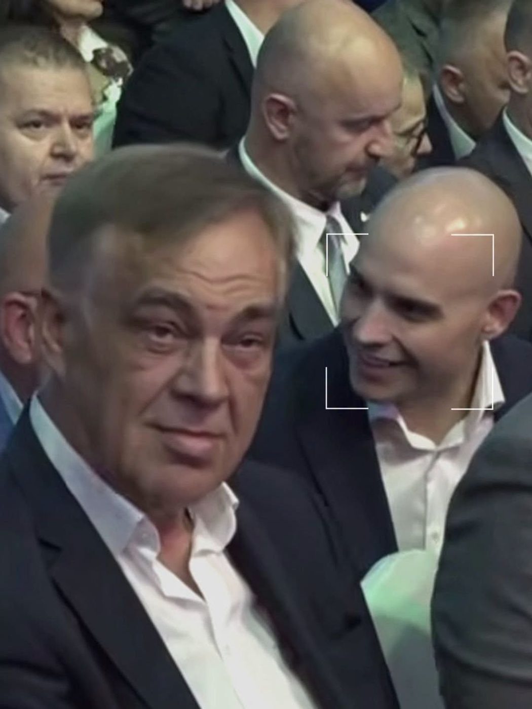 Video frame of a crowded indoor event with men in suits seated close together